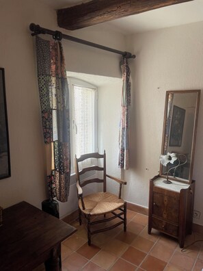 Ein Zimmer mit einem Holzstuhl, einem Holzschrank mit Spiegel und einem Fenster mit gemusterten Vorhängen.