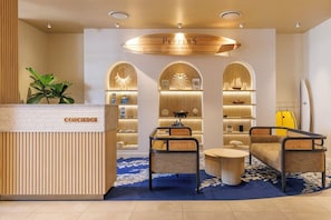 Zona de recepción de un hotel con un mostrador de madera identificado como 'CONCIERGE', una sala de estar con mobiliario de ratán y una tabla de surf apoyada en la pared.