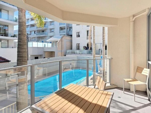 Un balcón con piscina, sillones de madera y vistas a edificios cercanos.