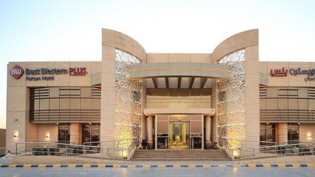L'ingresso di un hotel dal design moderno, caratterizzato da un arco centrale e due ali con le insegne 'Best Western PLUS' e 'Fursan Hotel'.
