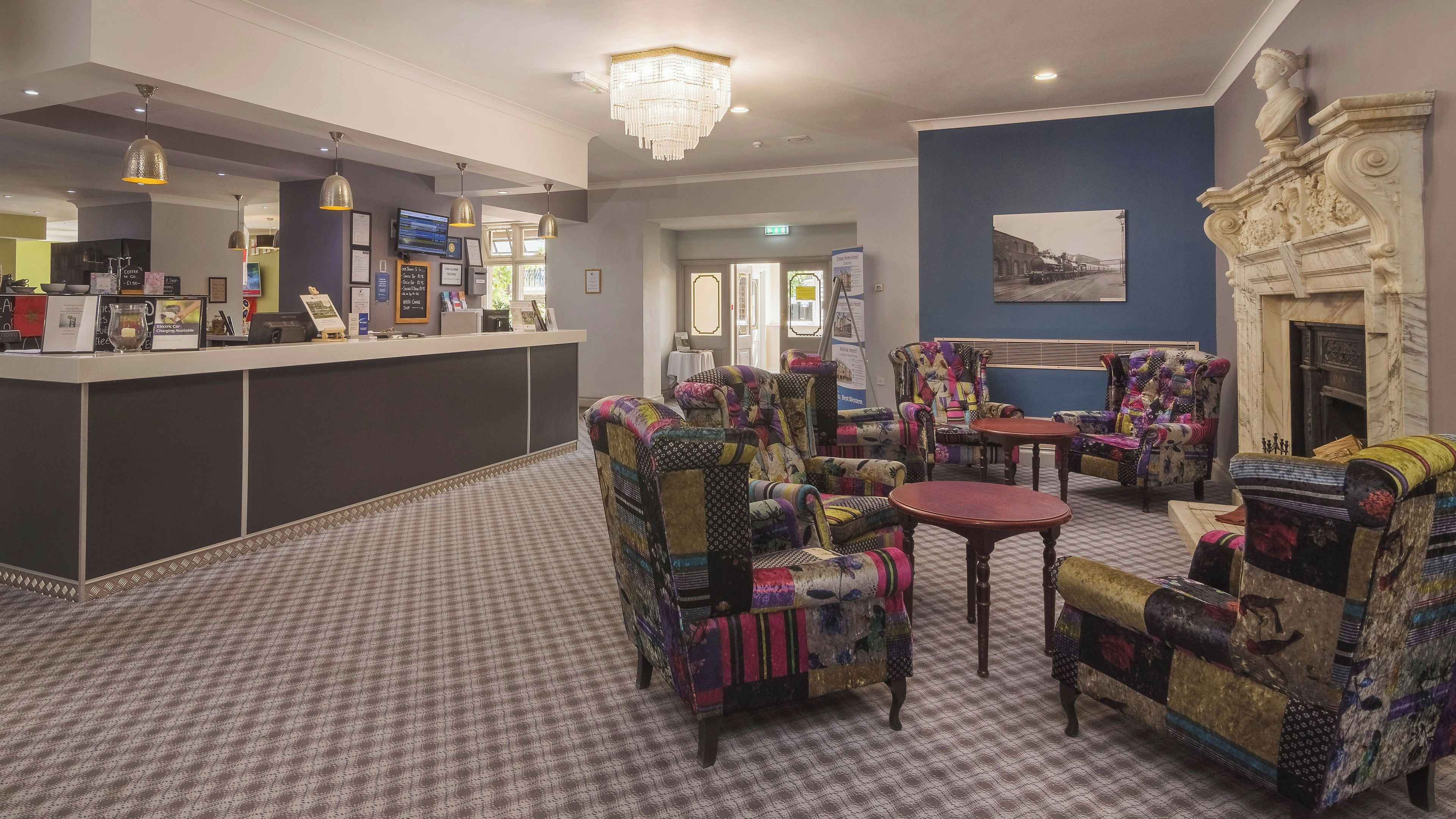 A hotel lobby with a reception desk, seating area with patterned chairs, a chandelier, and a fireplace.