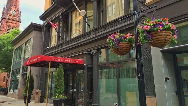 Un hôtel avec un auvent rouge, des jardinières suspendues remplies de fleurs et, en arrière-plan, un clocher.
