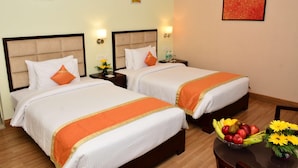 A hotel room with two beds, a nightstand with a lamp, a table with fruit and flowers, and artwork on the walls. - Sagar Sona (Lucknow)