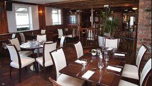 A restaurant with wooden tables and chairs, a brick wall, and a large window. - The White Sands Hotel Portmarnock (Portmarnock)