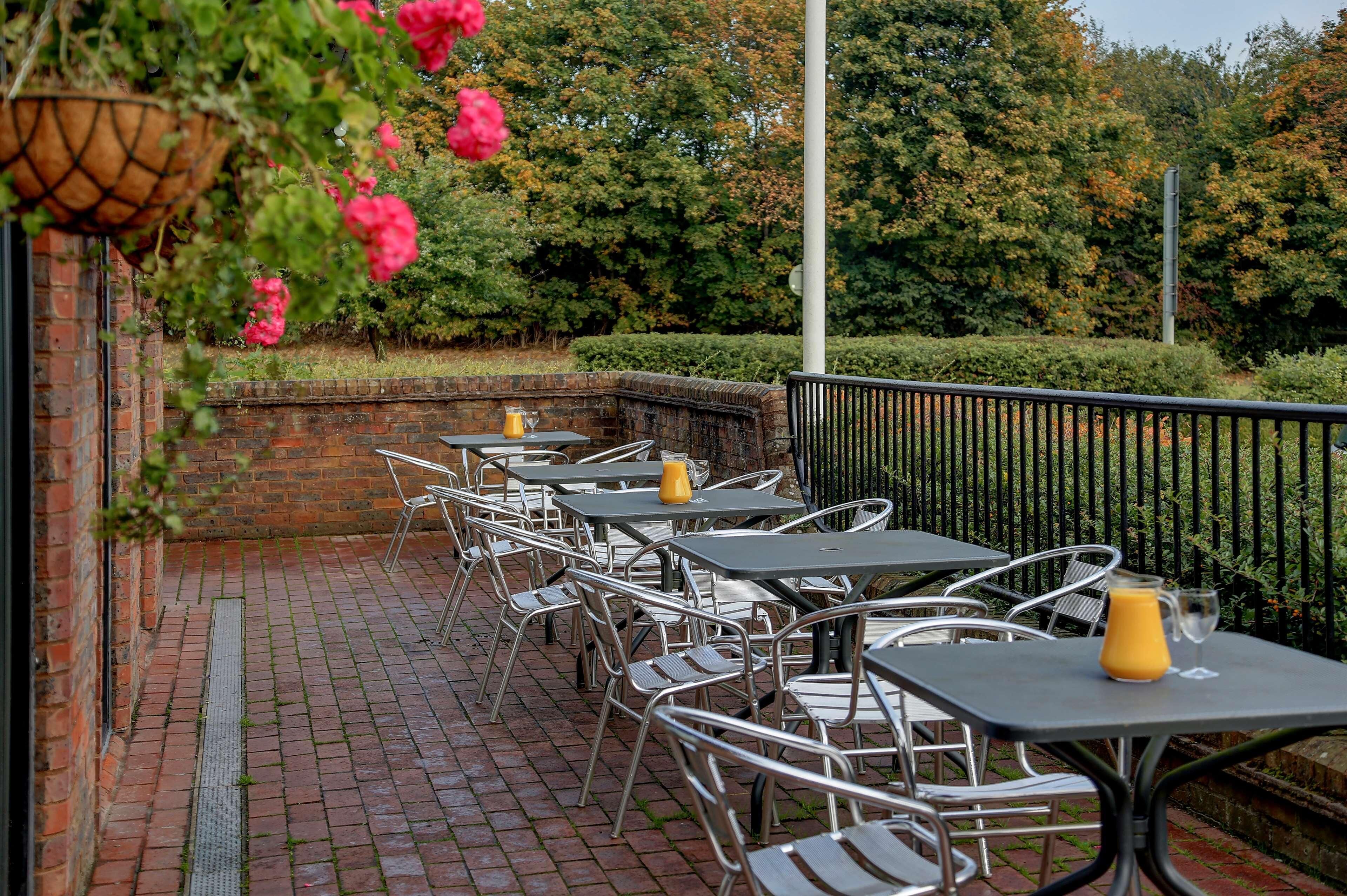 An outdoor seating area with tables and chairs, a brick wall, and a hedge in the background.