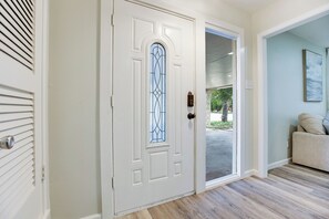 Une porte blanche avec un panneau de verre et un trou de serrure, qui donne sur un couloir au sol de couleur claire et où se trouve un canapé beige.