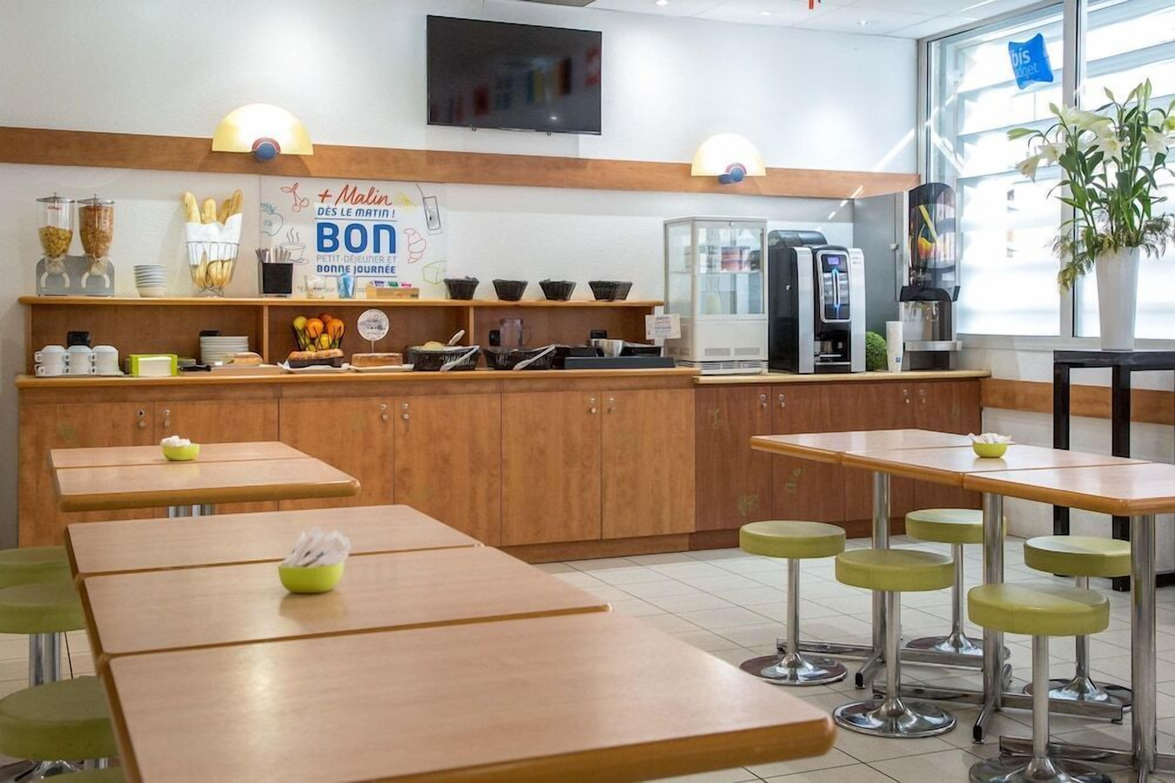 Un café avec des tables en bois et des tabourets verts, une machine à café et une enseigne sur laquelle on peut lire : « Malin Bonjour ».