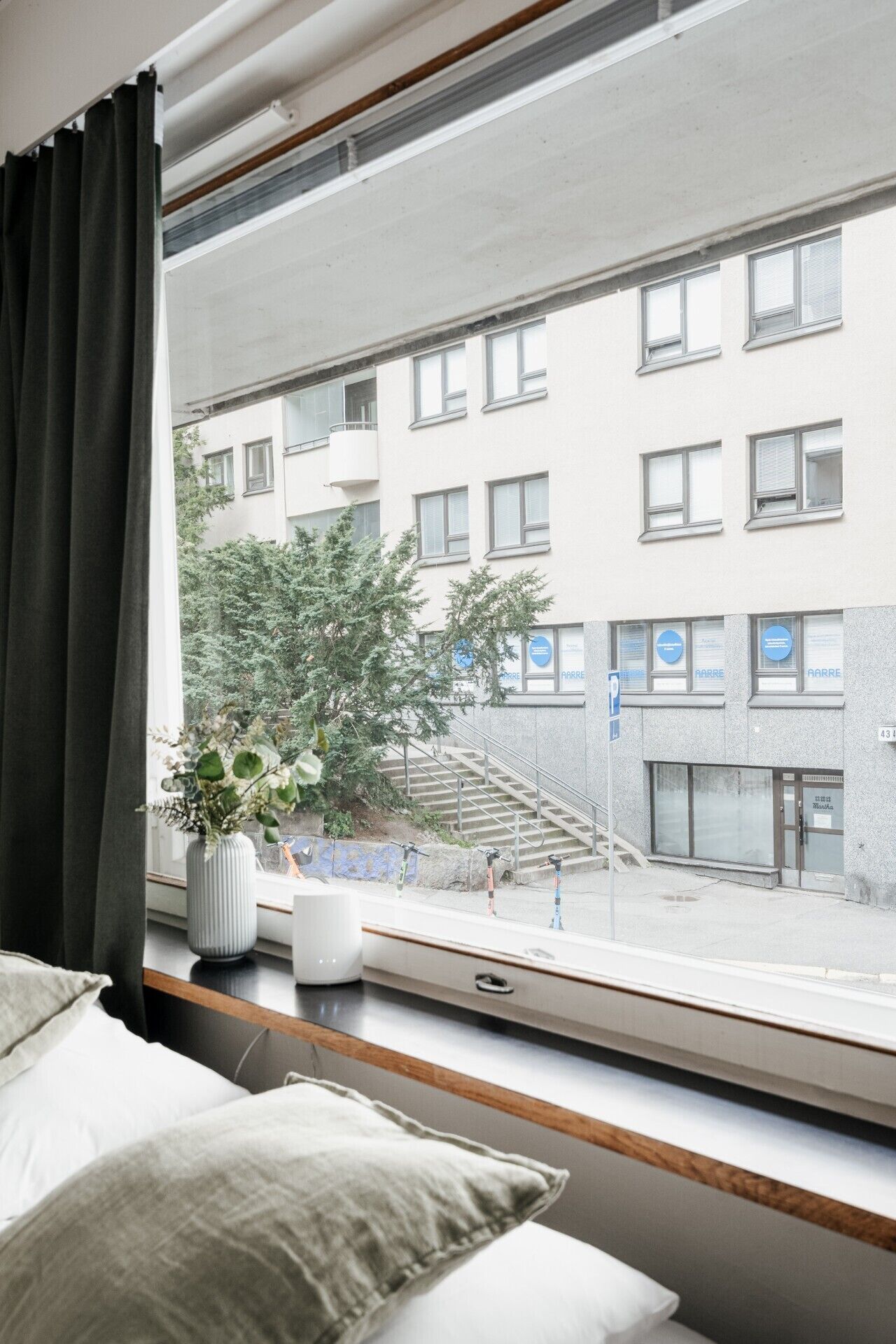 Une pièce avec une fenêtre donnant sur un immeuble doté de nombreuses fenêtres et d’un escalier. On aperçoit une plante sur le rebord de la fenêtre et un lit avec des oreillers.