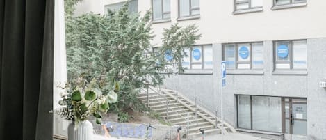 A room with a window overlooking a building with multiple windows and a staircase. There is a plant on the windowsill and a bed with pillows.