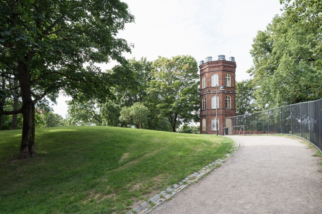 A brick tower structure with a pathway leading to it, surrounded by trees and a fenced area.