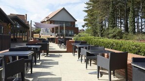 An outdoor patio area with tables and chairs, a building with a sloped roof, and a backdrop of trees. - Le Strange Arms Beachside Hotel & Lodges (Hunstanton)