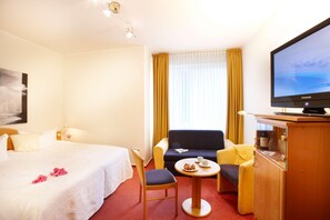 Une chambre d’hôtel avec un lit, un canapé, une petite table avec une bouteille de vin et des verres, une télévision et une fenêtre avec des rideaux.