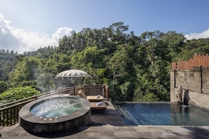 A hot tub with a wooden deck and lounge chairs, surrounded by lush greenery and a clear sky.