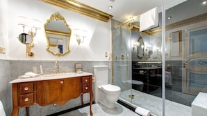 Un baño lujoso con suelo de mármol, tocador de madera con detalles dorados, inodoro blanco, ducha con mampara de vidrio y un espejo grande con adornos dorados.