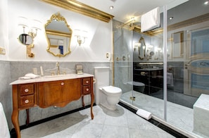 Un baño lujoso con suelo de mármol, tocador de madera con detalles dorados, inodoro blanco, ducha con mampara de vidrio y un espejo grande con adornos dorados.