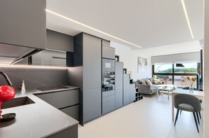 A modern kitchen with integrated appliances, a dining area with a view, and a living space with a sofa and table. - GRAN RESERVA MIMOSES ATIC B (Empuriabrava)