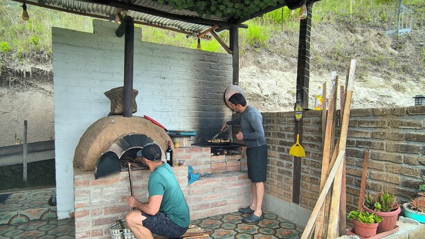 Una cocina rústica al aire libre con horno de ladrillo, suelo de baldosas y un porche con plantas colgantes.