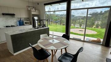 A modern kitchen with a large island, a dining area with a table and chairs, and a view of a garden with a hammock and a dog.