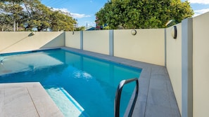 A swimming pool with a clear blue water, surrounded by a light-colored wall and a tiled deck.