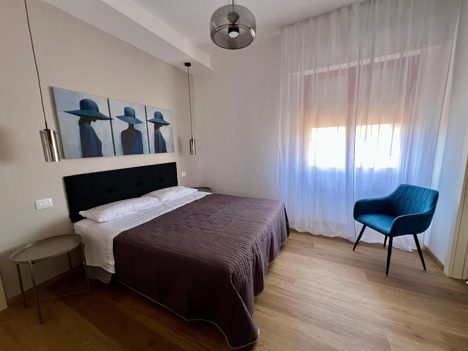 A hotel room with a bed, a chair, a painting on the wall, and a window with curtains.