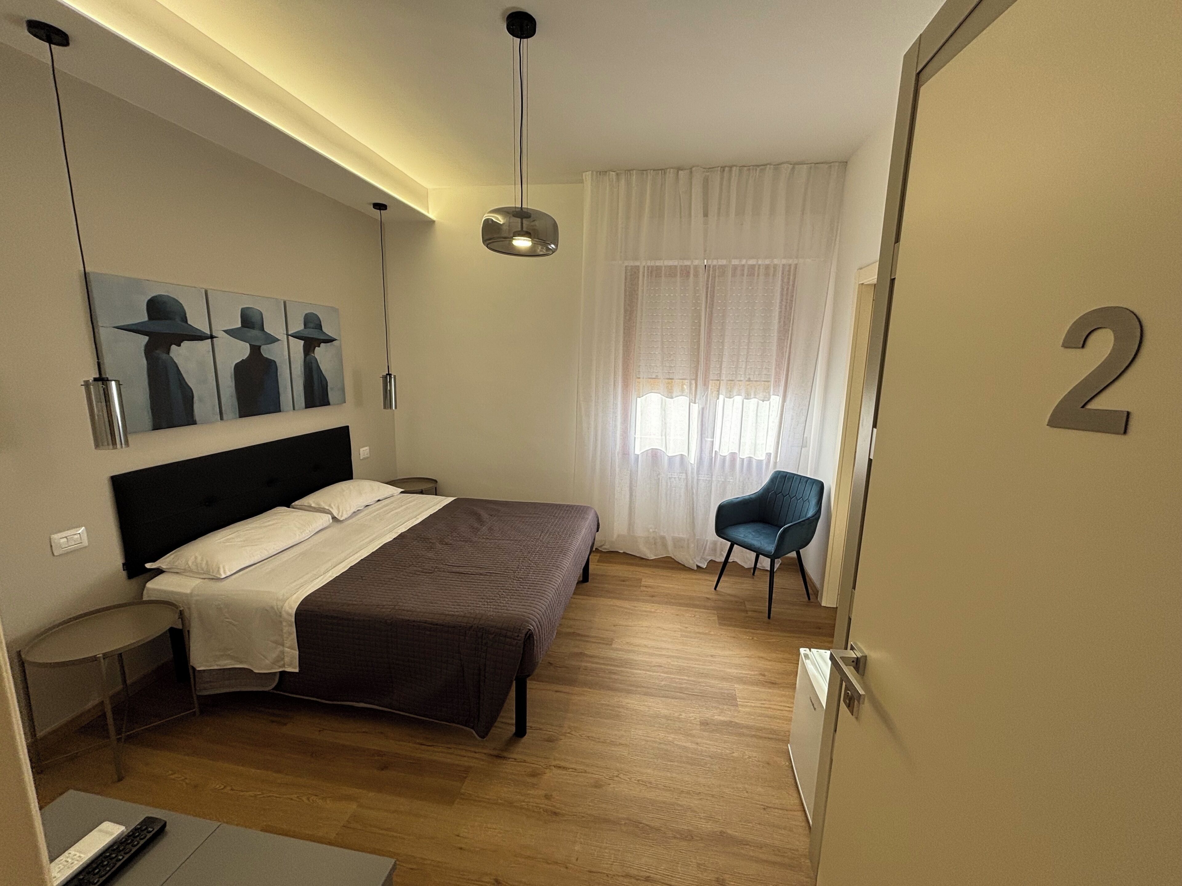 Un dormitorio con cama, una silla, un cuadro, una ventana con cortinas y una puerta con un número.