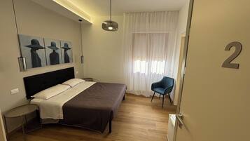 Un dormitorio con cama, una silla, un cuadro, una ventana con cortinas y una puerta con un número.