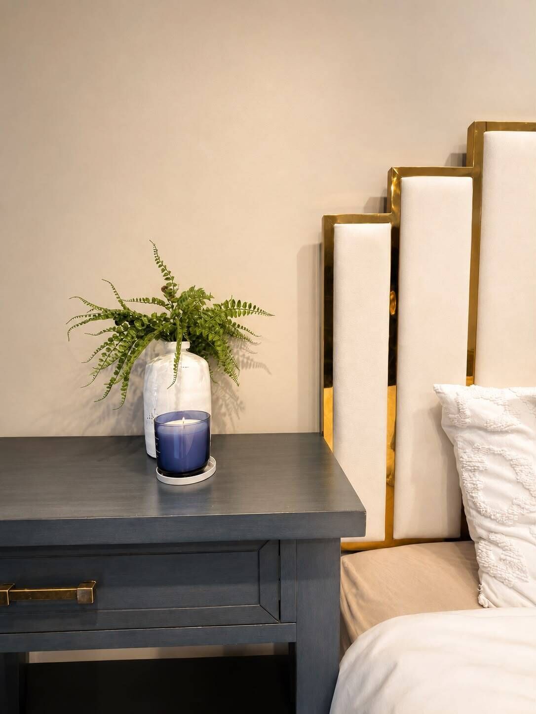 A bedside table with a blue candle, a white vase with a green plant, and a bed with white bedding and a gold-framed headboard.