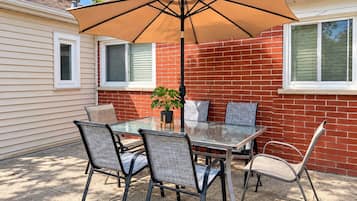 Une terrasse extérieure avec une table en verre, quatre chaises, un pot de fleurs et un grand parasol.