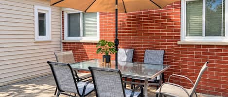 An outdoor patio with a glass table, four chairs, a potted plant, and a large umbrella.