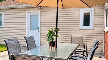 An outdoor patio with a glass table and chairs, a large umbrella, and a potted plant.