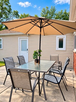 An outdoor patio with a glass table and chairs, a large umbrella, and a potted plant.