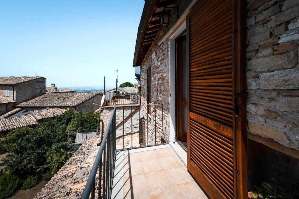 Ein Steinhaus mit einer hölzernen, verstellbaren Tür, einem Balkon mit Metallgeländer und Blick auf terrassenförmig angeordnete Häuser.