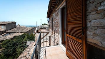 Ein Steinhaus mit einer hölzernen, verstellbaren Tür, einem Balkon mit Metallgeländer und Blick auf terrassenförmig angeordnete Häuser.