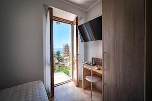 Un dormitorio con cama, escritorio y armario. Tiene un balcón con vistas a edificios.