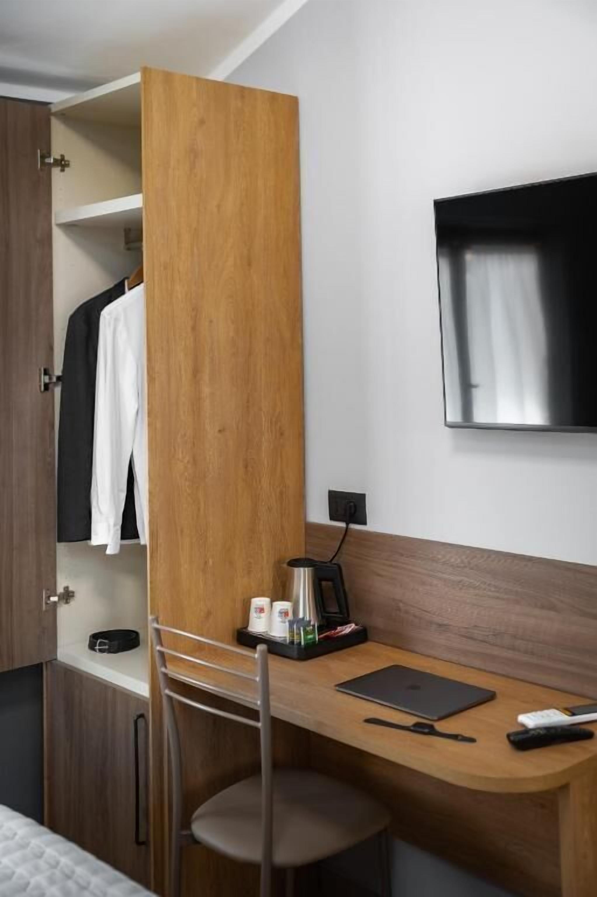 Ein Hotelzimmer mit einem Holztisch, einem Sessel, einem Bett, einem an der Wand befestigten Fernseher und einem Kleiderschrank.
