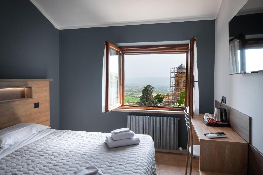 A hotel room with a bed, a desk, a chair, a window with a view of a building, and a radiator.