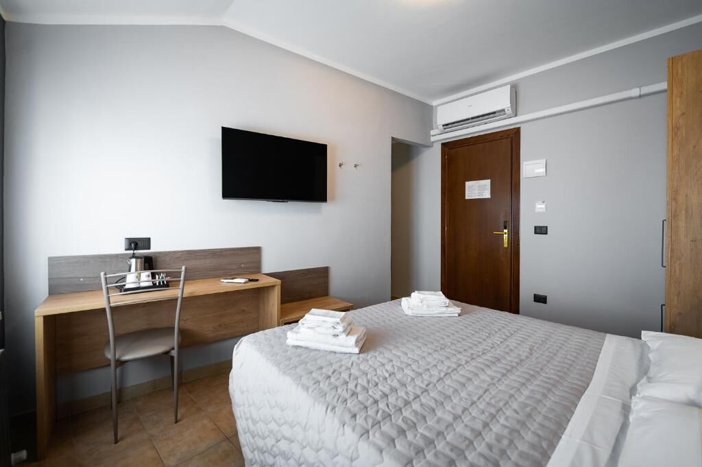 Une chambre d’hôtel comprenant un lit, un bureau avec une chaise, un téléviseur à écran plat, un appareil de climatisation et une porte en bois.