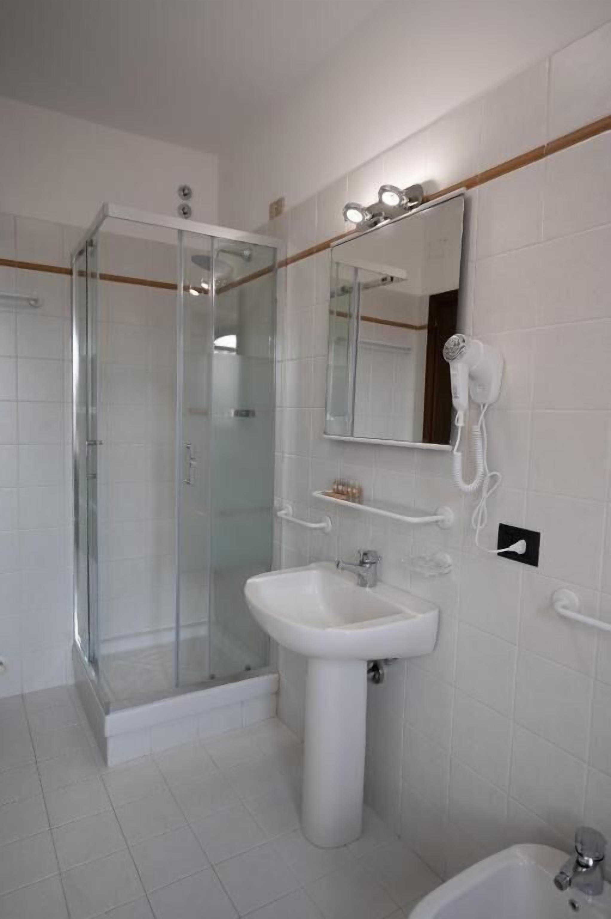 Casa de banho com chuveiro em cabine de vidro, lavatório com coluna, espelho e secador de cabelo fixado na parede.