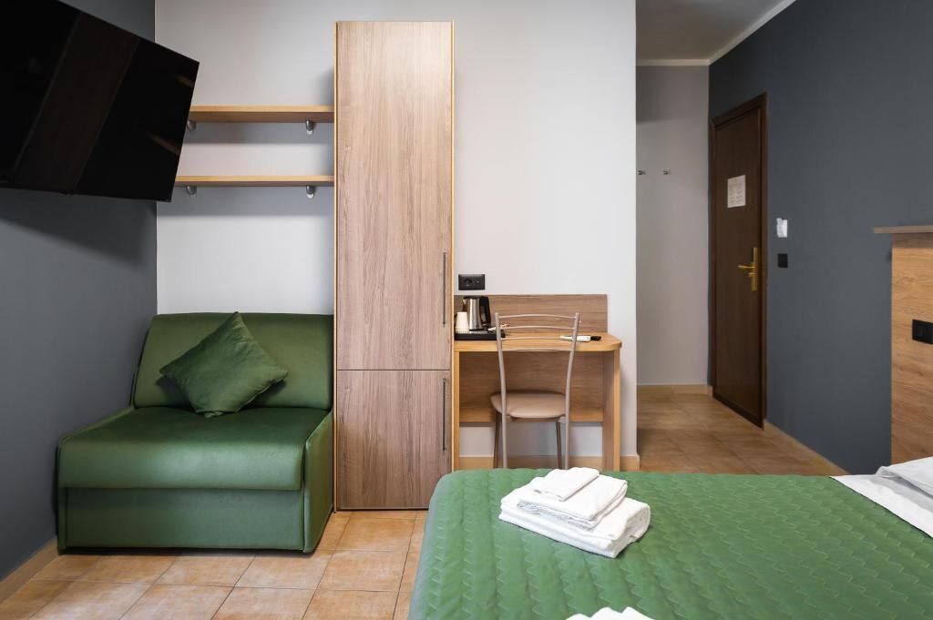A compact room with a green sofa, a bed with a green bedspread, a wooden desk with a chair, and a television mounted on the wall.