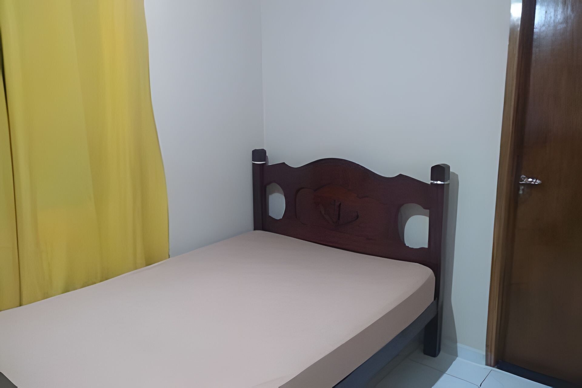 Una cama individual con cabecera de madera en una habitación con puerta y cortina amarilla.