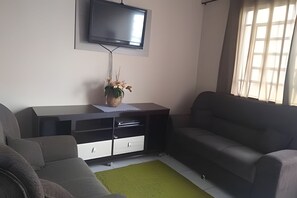 A living room with a flat-screen TV mounted on the wall, a dark-colored sofa, a white cabinet with drawers, and a green rug on the floor. - Casa do Bruno (Zangirolami)