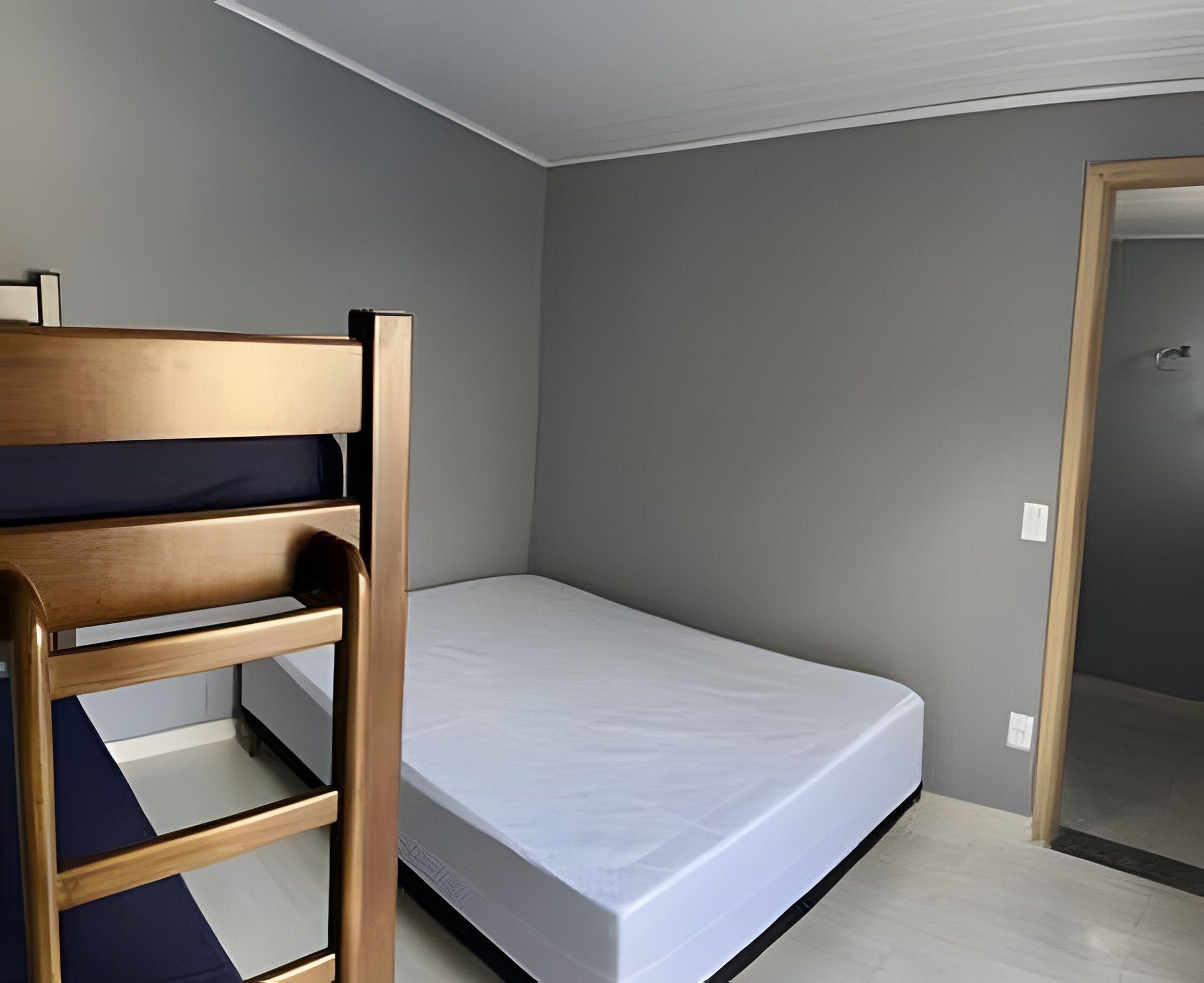 A small, single-bed room with a wooden bunk bed on one side and a plain mattress on the other.