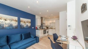 A modern living room with a blue sofa, a dining area with a table and chairs, a kitchen with white cabinets, and two abstract paintings on the wall. - Ibex House Cozy Modern 2-bed Flat Free Parking (Reading)