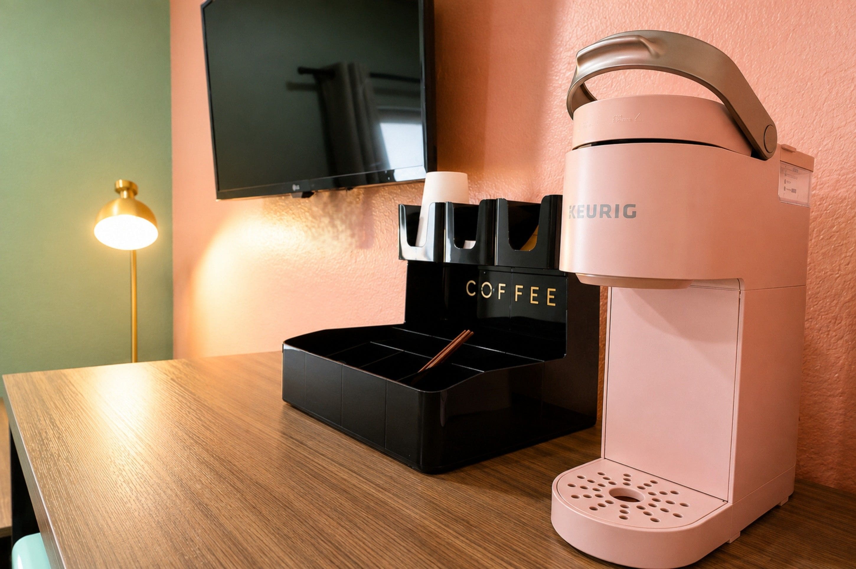 Una cafetera con un soporte para cápsulas de café sobre una mesa de madera, una lámpara y un televisor de pantalla plana en la pared.
