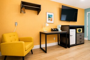 A room with a yellow armchair, a small table with a lamp, a television, a mini-fridge, and a shelf with hanging items.