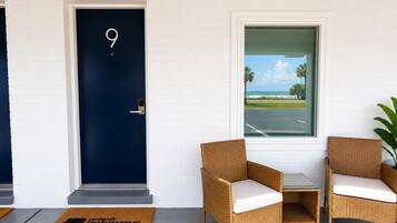 Une pièce avec une porte bleu foncé portant le numéro 9, des chaises en osier avec des coussins blancs, une petite table et une vue sur une plage, visible par la fenêtre.