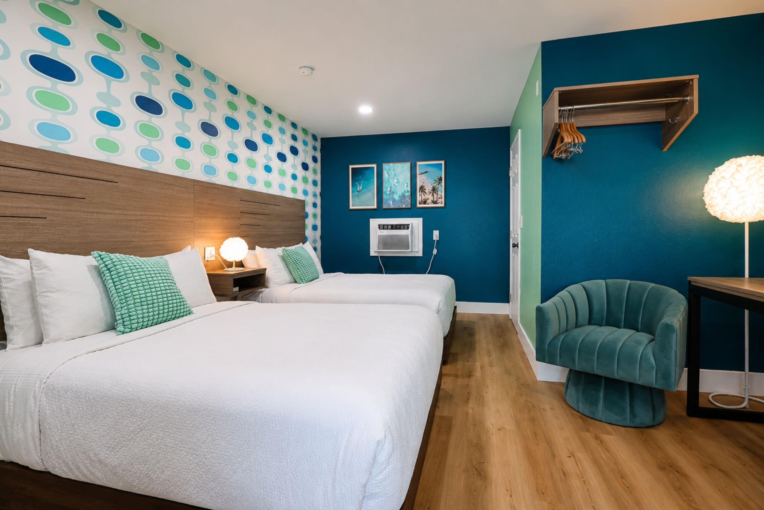 Ein Hotelzimmer mit zwei Betten, einem Holzkopfteil, einer Wand-Klimaanlage und einem petrolfarbenen Sessel.
