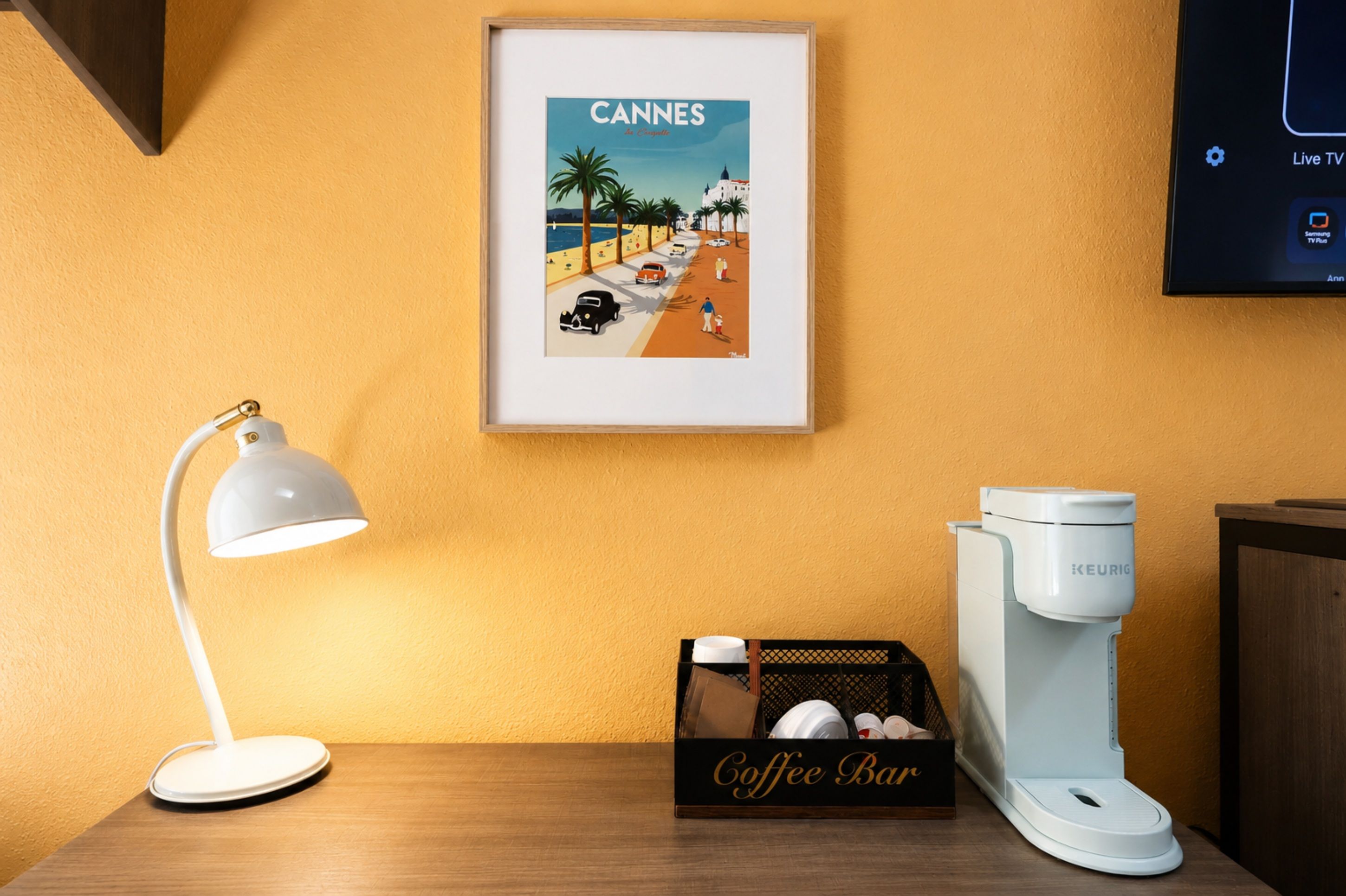 Une photo encadrée représentant une scène de plage, avec le mot « CANNES » écrit sur le mur, une lampe de bureau blanche, une machine à café et une boîte étiquetée « Bar à café ».