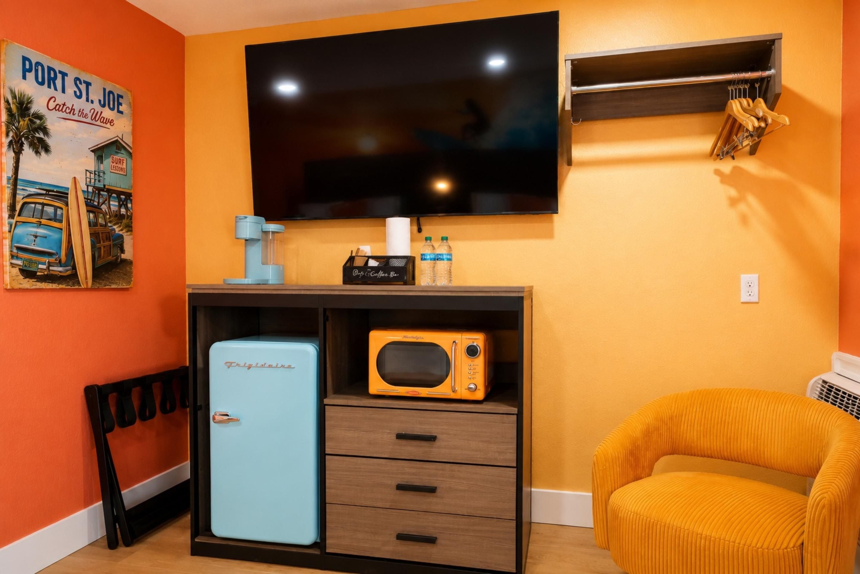A room with a television mounted on the wall, a small refrigerator, a microwave, a chair, and a shelf with a surfboard and a sign.