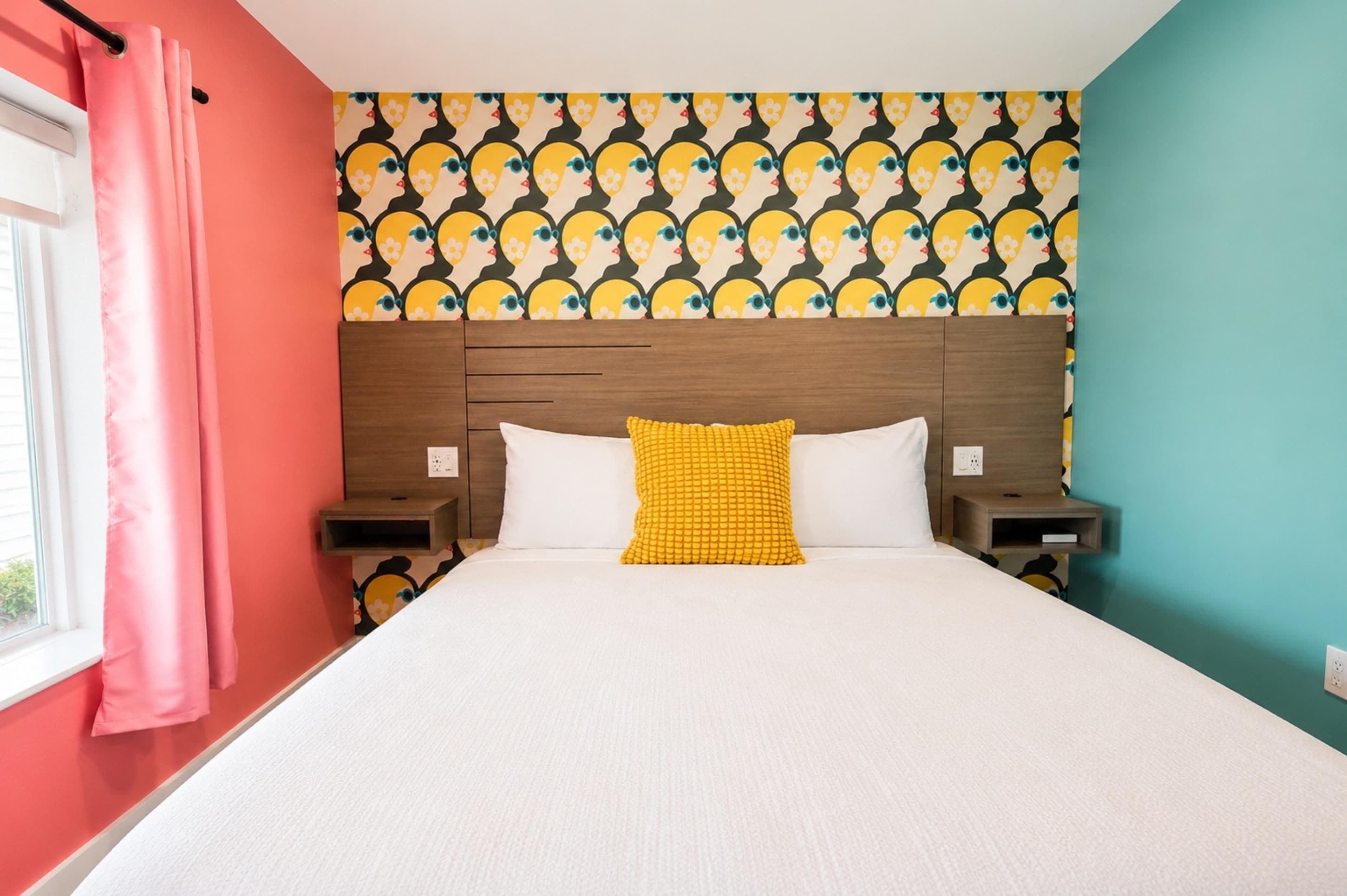 Habitación de hotel con cama, cabecero, dos mesitas de noche, ventana con cortinas y un papel tapiz estampado.